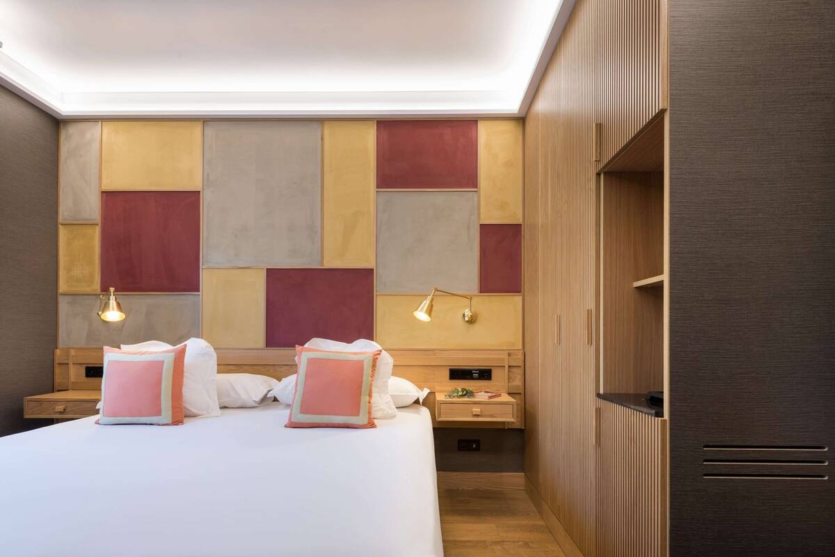 A modern hotel room with a large bed, bedside tables, a wooden wardrobe, and a wall with geometric patterns.