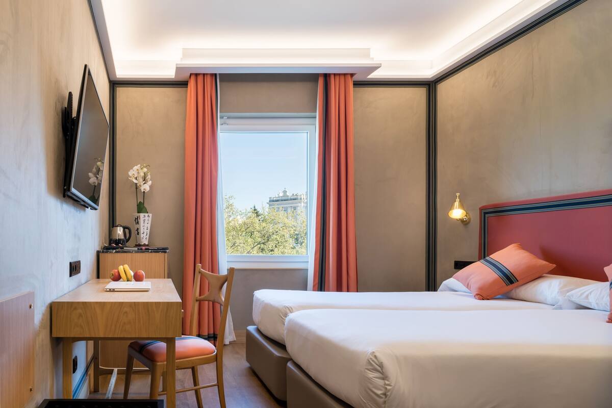 A hotel room with a bed, a desk with a laptop, a chair, a window with curtains, and a view of a building outside.