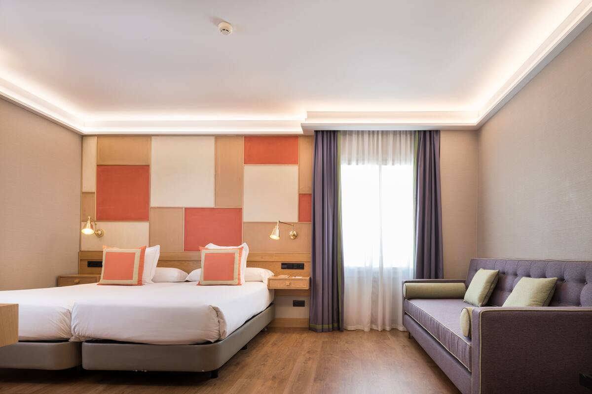 A hotel room with a double bed, a sofa, a nightstand, and a window with curtains.