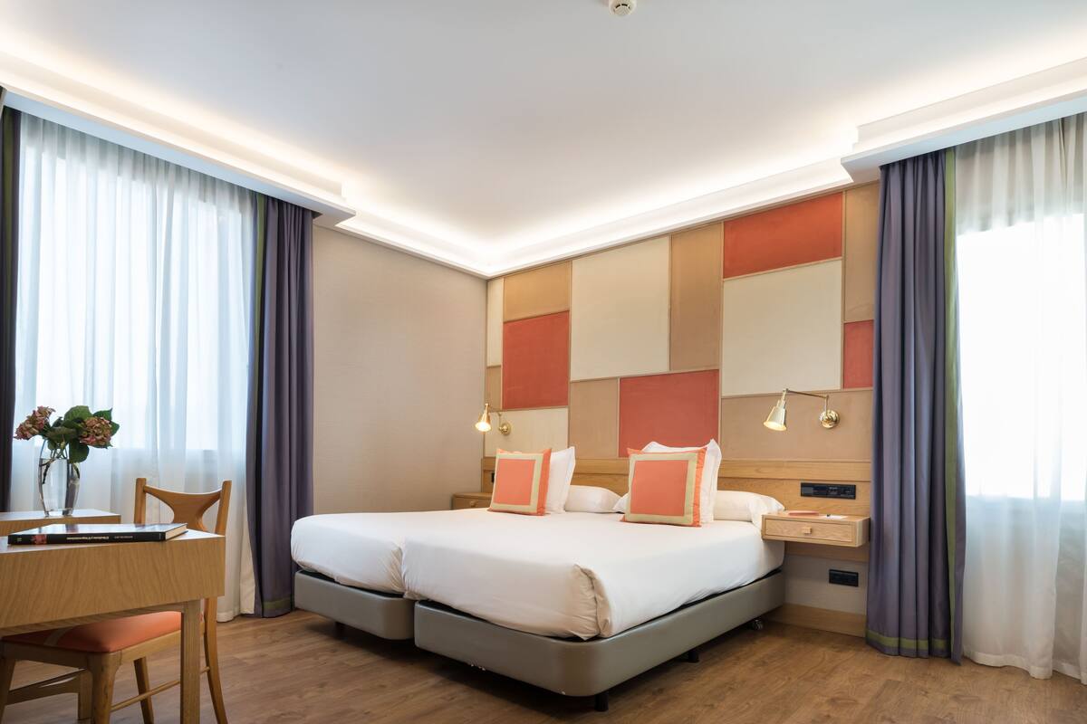 A hotel room with a double bed, bedside tables, a desk with a chair, and a large window with curtains.
