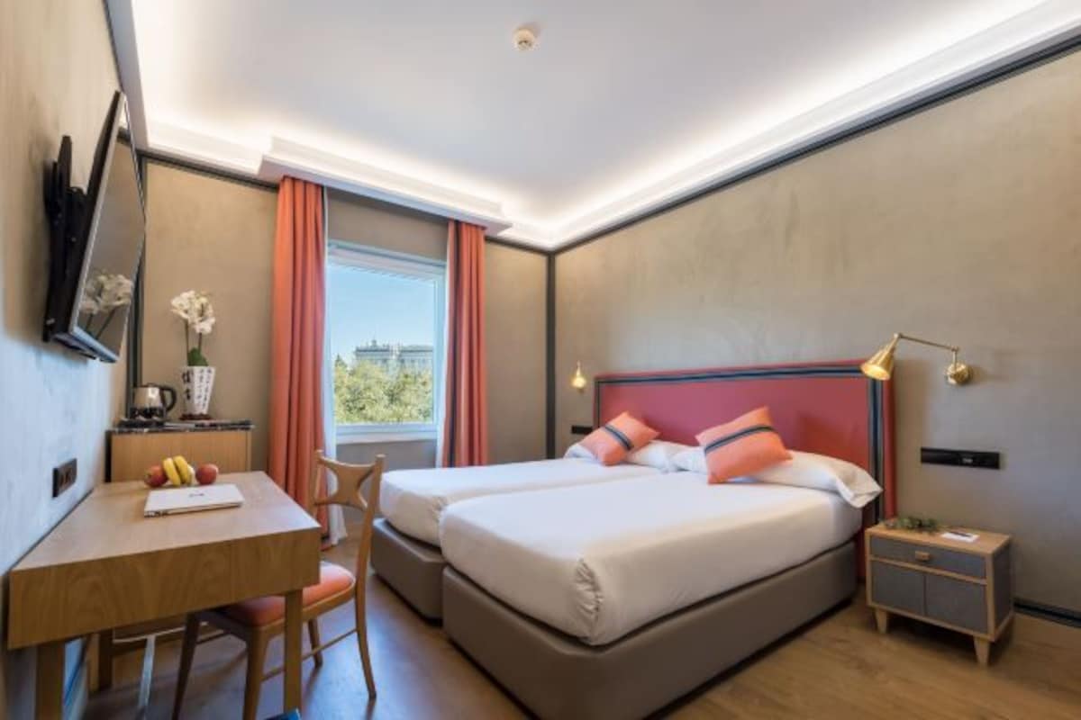 Una habitación de hotel moderna con una cama grande, un escritorio con una silla, una mesita con frutas, una mesita de noche y una ventana con cortinas.
