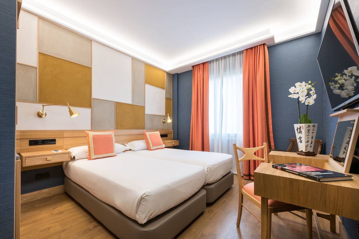 A hotel room with two beds, a desk, a chair, a bedside table, a lamp, a vase with flowers, and a large mirror.