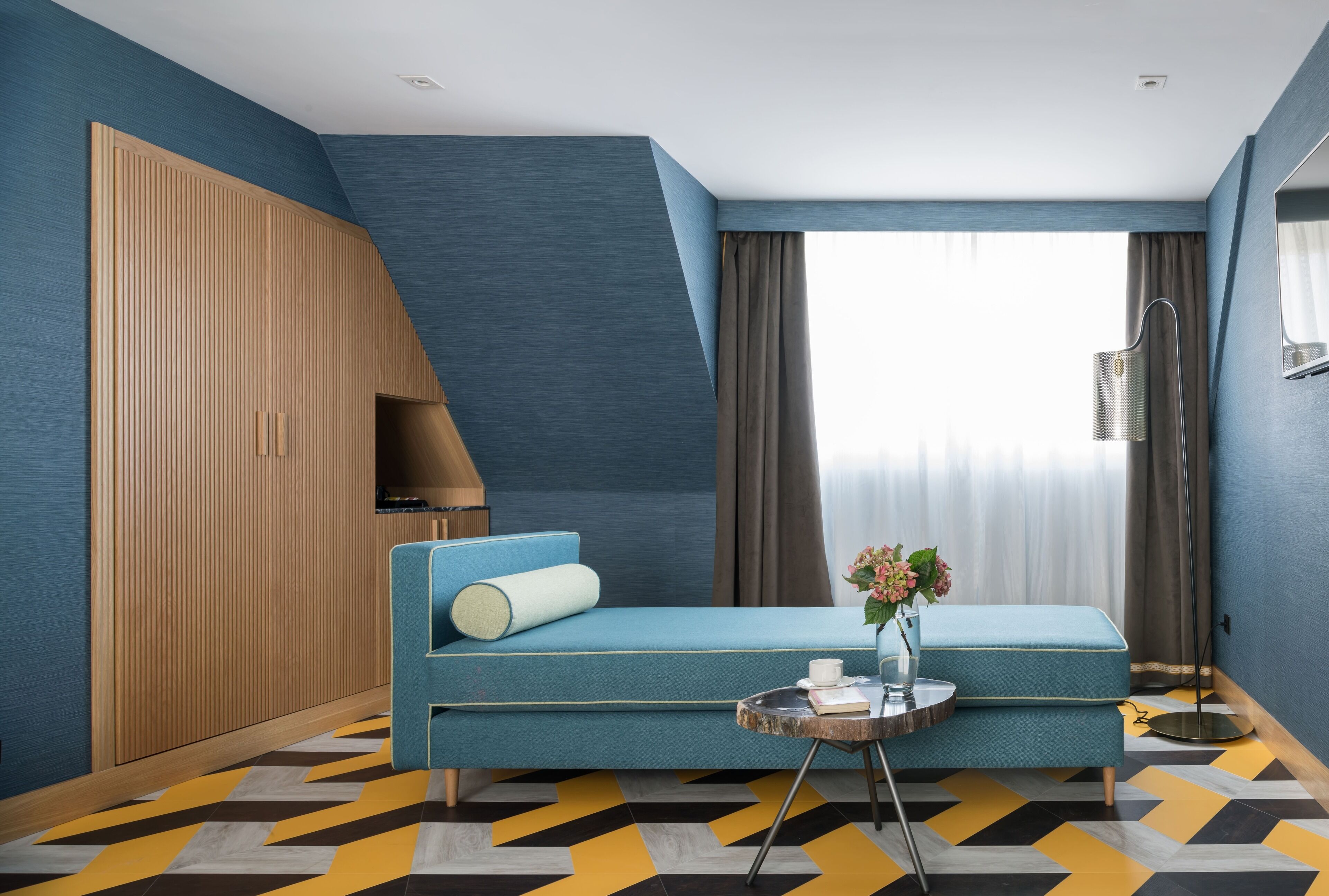 Una habitación de hotel moderna con un sofá azul, un armario de madera, una mesita con un florero y un suelo con estampado geométrico.