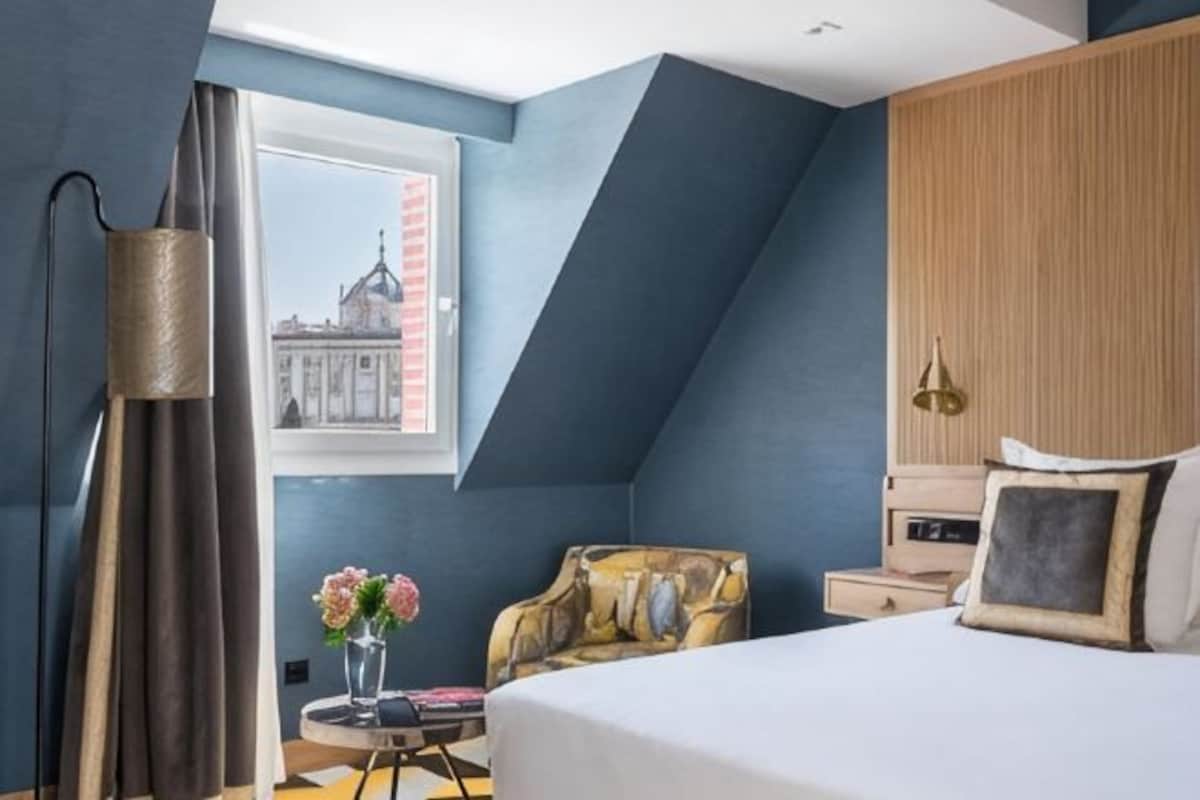 Un dormitorio con cama, una silla, una mesita de noche, una lámpara, una ventana con vistas a un edificio y un florero con flores.