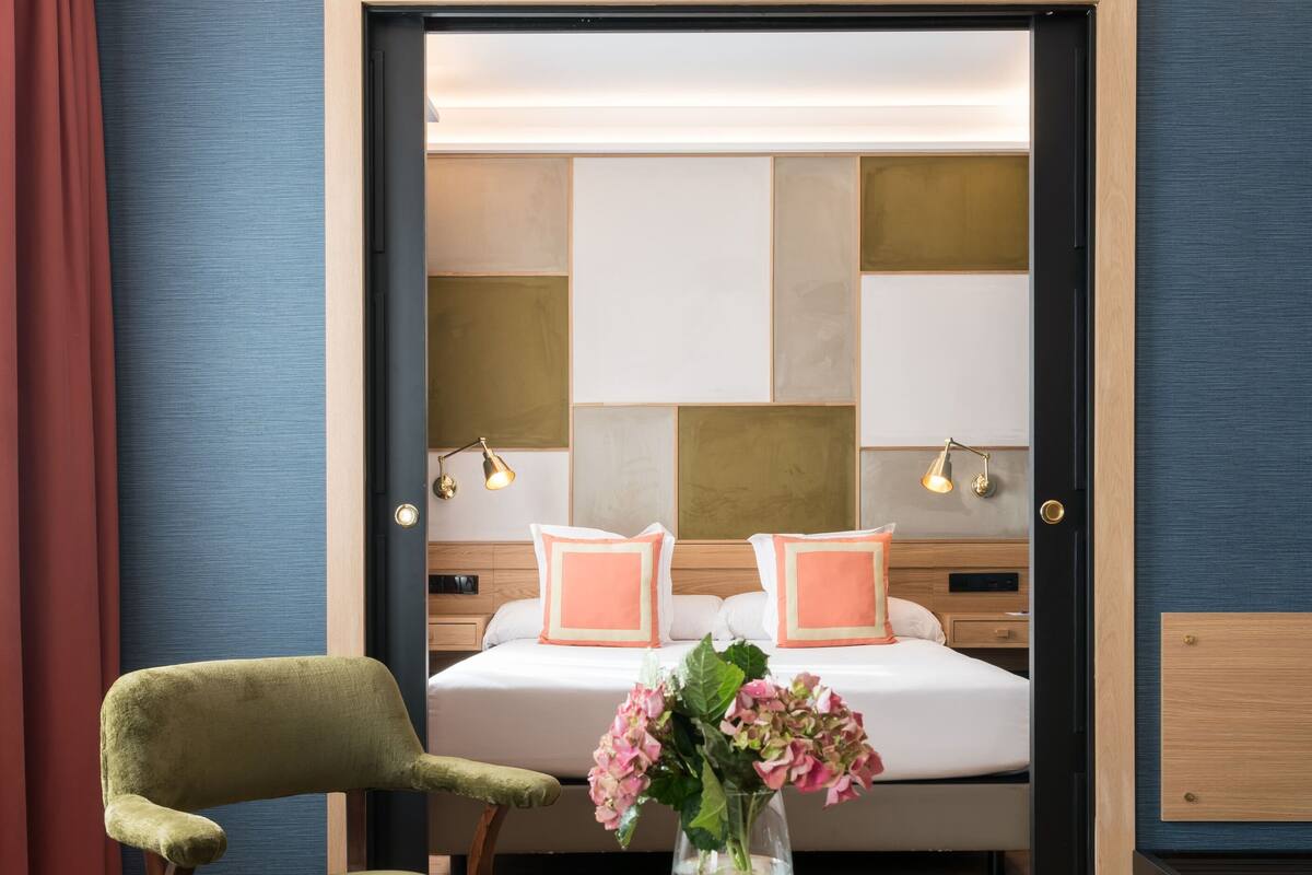 A hotel room with a bed, a green chair, a glass table, and a vase with flowers.