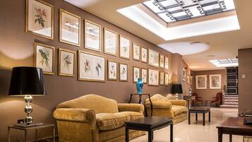 Eine Hotelhalle mit hoher Decke, Oberlichtern, gerahmten Kunstwerken, Sofas, Tischen und einer Treppe.