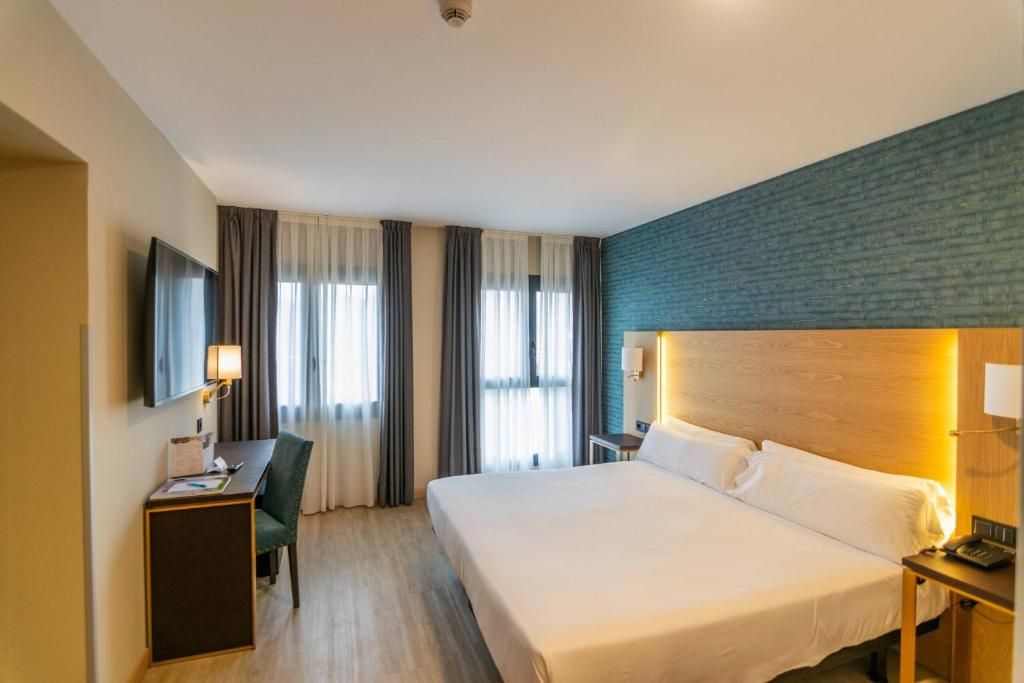 A hotel room with a bed, a desk with a chair, a television, and a nightstand with a lamp.