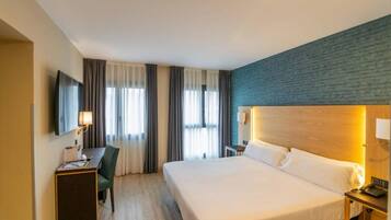 A hotel room with a bed, a desk with a chair, a television, and a nightstand with a lamp.