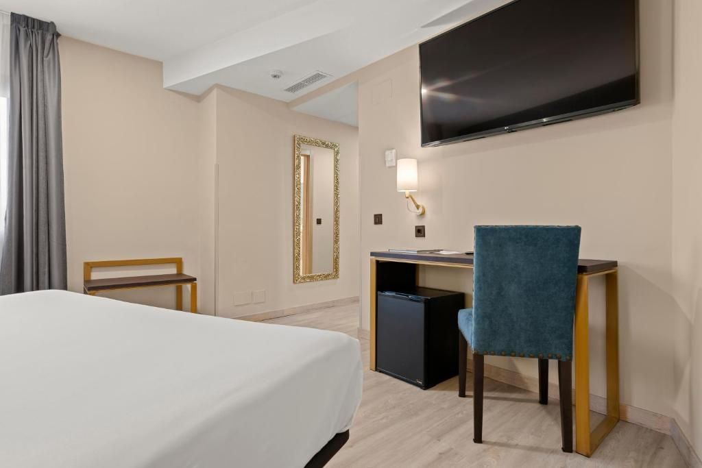 A hotel room with a bed, desk, chair, TV, and a small refrigerator.