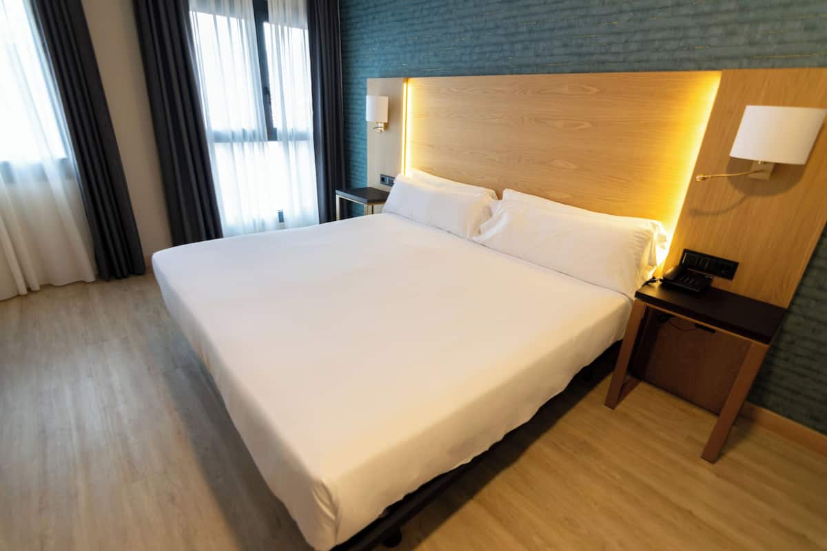 Un grand lit avec du linge de lit blanc, une tête de lit en bois et une table de chevet avec une lampe.