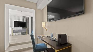 A hotel room with a flat-screen TV mounted on the wall, a desk with a lamp, and a chair.