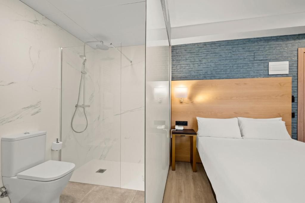 A hotel room with a shower, toilet, bed, bedside table, and a wall-mounted light.