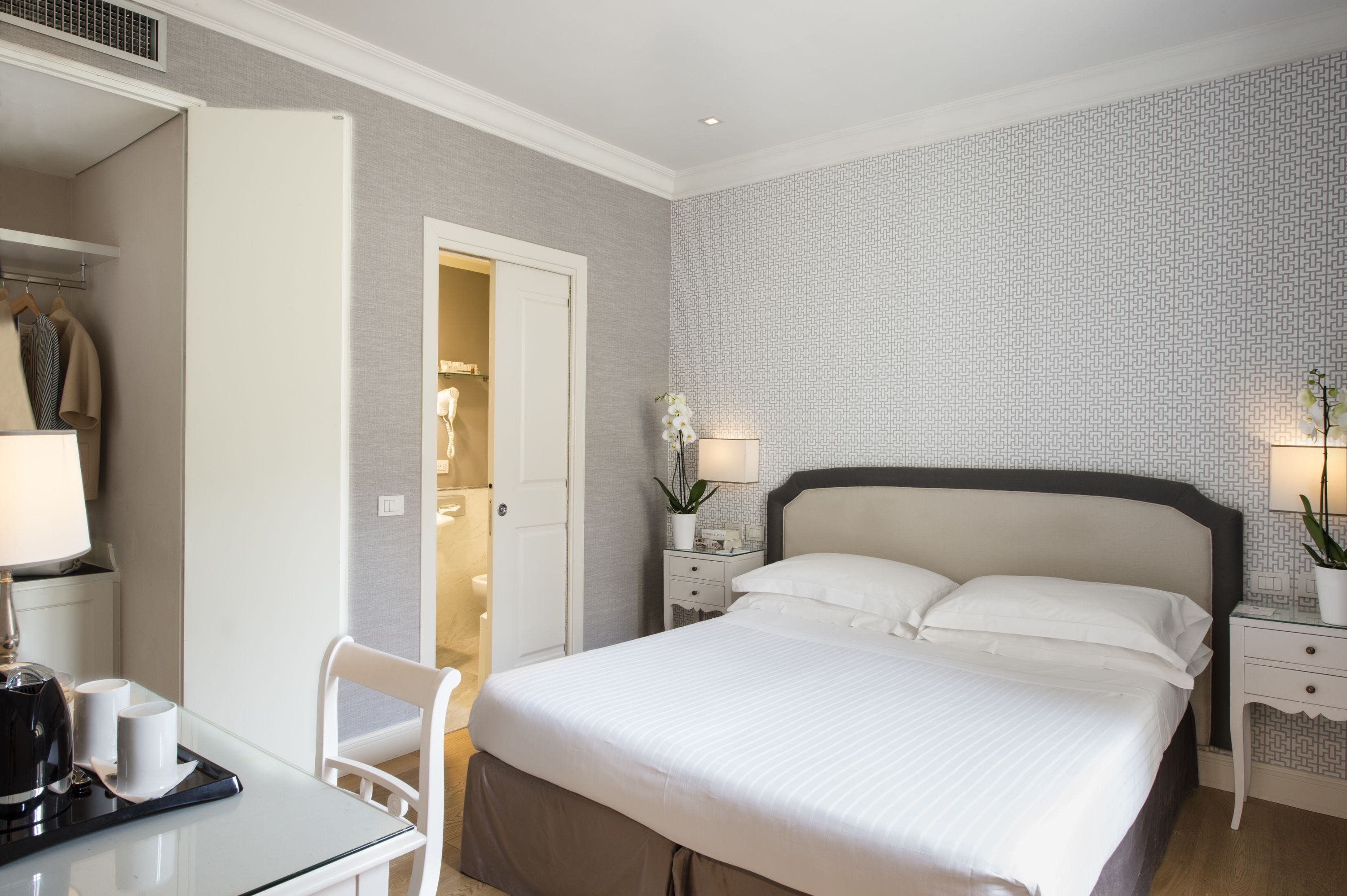 A neatly arranged hotel room with a large bed, a bedside table with a lamp, a chair, a desk with a coffee maker, and a wardrobe.