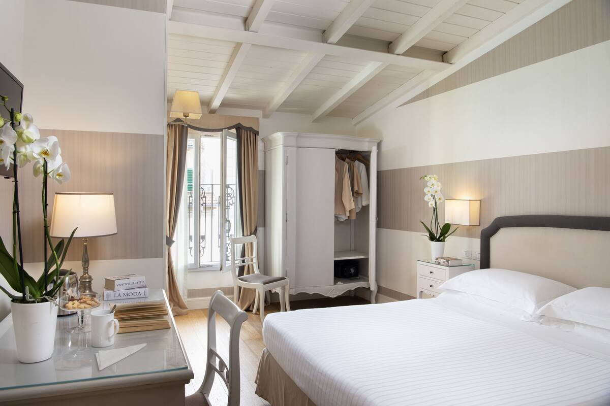 A hotel room with a bed, a desk with a lamp, a chair, a wardrobe, and a potted orchid.