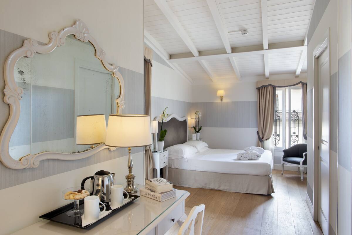A hotel room with a large ornate mirror, a bed with white linens, a bedside table with a lamp, a tray with breakfast items, and a view of the balcony.