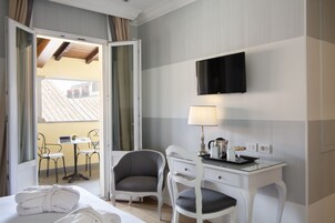 Une chambre d’hôtel comprenant un lit, deux chaises, un bureau avec une lampe, un téléviseur à écran plat et une porte ouverte donnant sur un balcon équipé d’une table et de chaises.