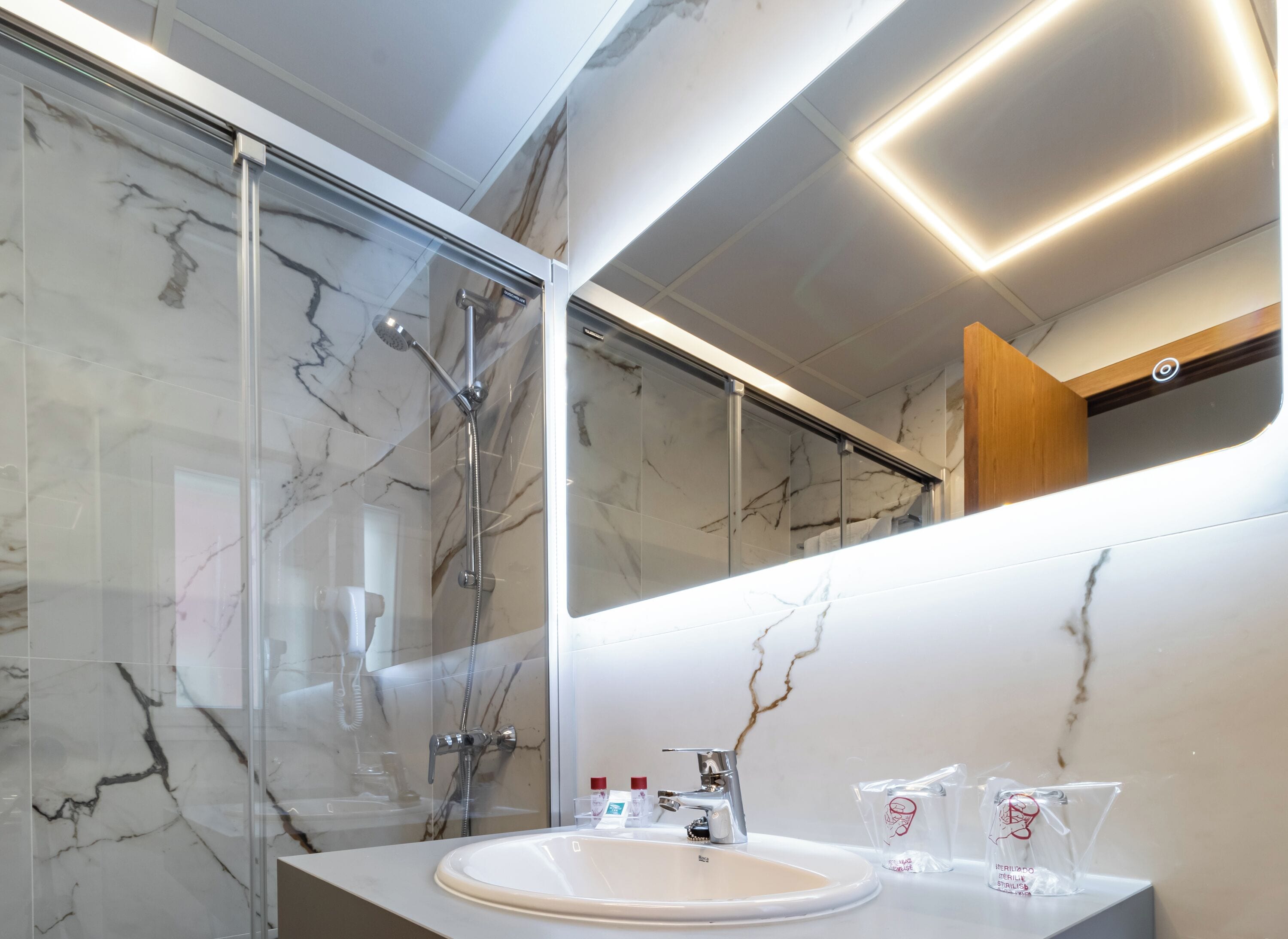 A modern bathroom with a glass shower enclosure, a marble-tiled wall, a white sink, and a mirror with integrated lighting.