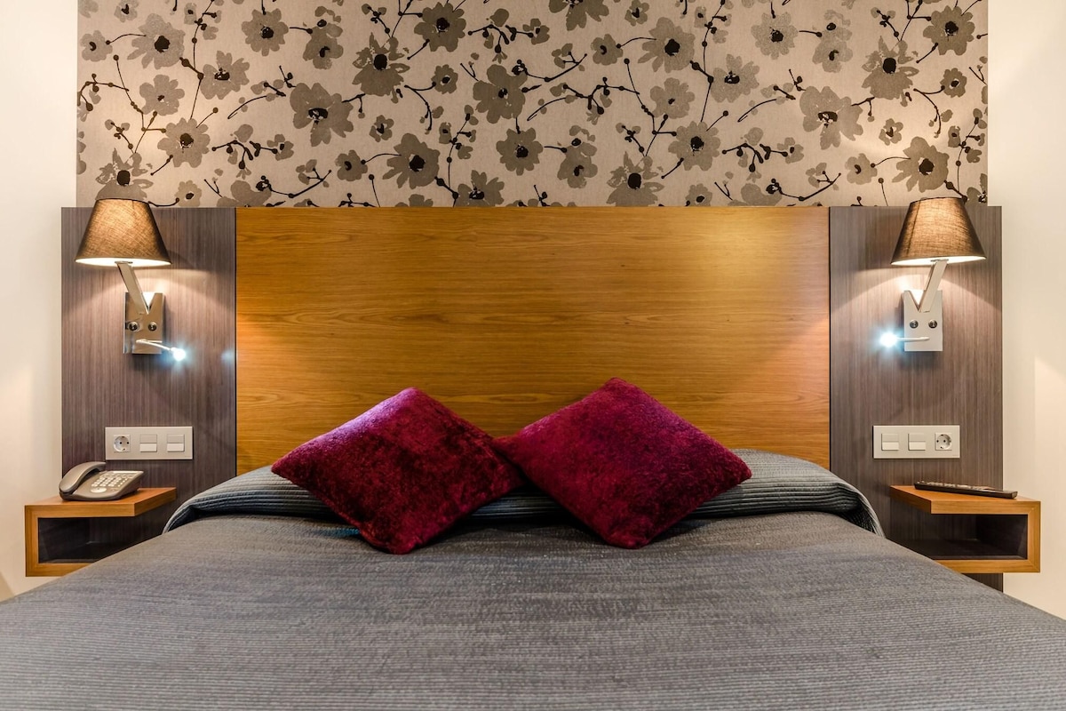 Una cama bien tendida con dos almohadas rojas, un cabecero de madera y dos lámparas de noche.