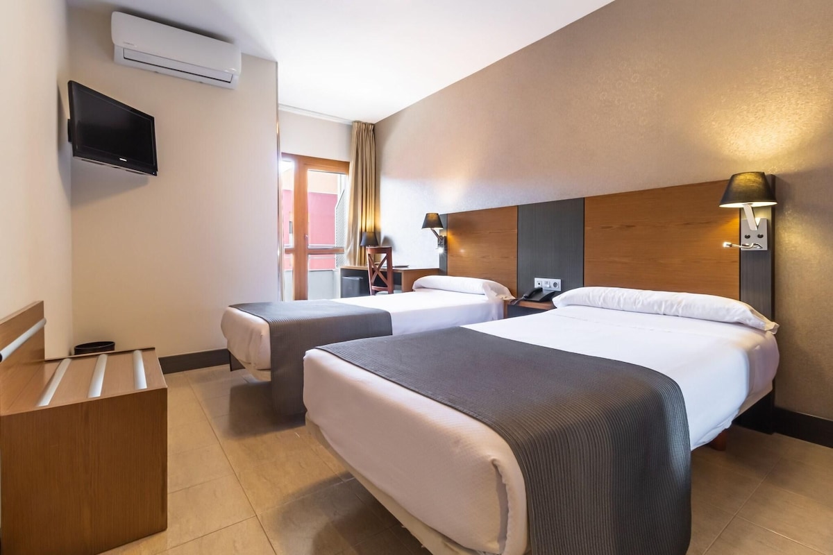 A hotel room with two beds, a desk, a TV, and a wall-mounted lamp.