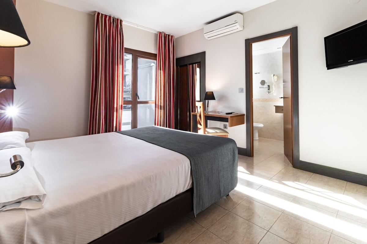 A hotel room with a bed, a desk, a chair, an air conditioner, a TV, and a bathroom visible through an open door.