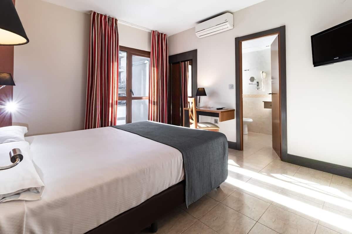 Habitación de hotel con cama, escritorio, silla, aire acondicionado, televisor y baño visible a través de una puerta abierta.