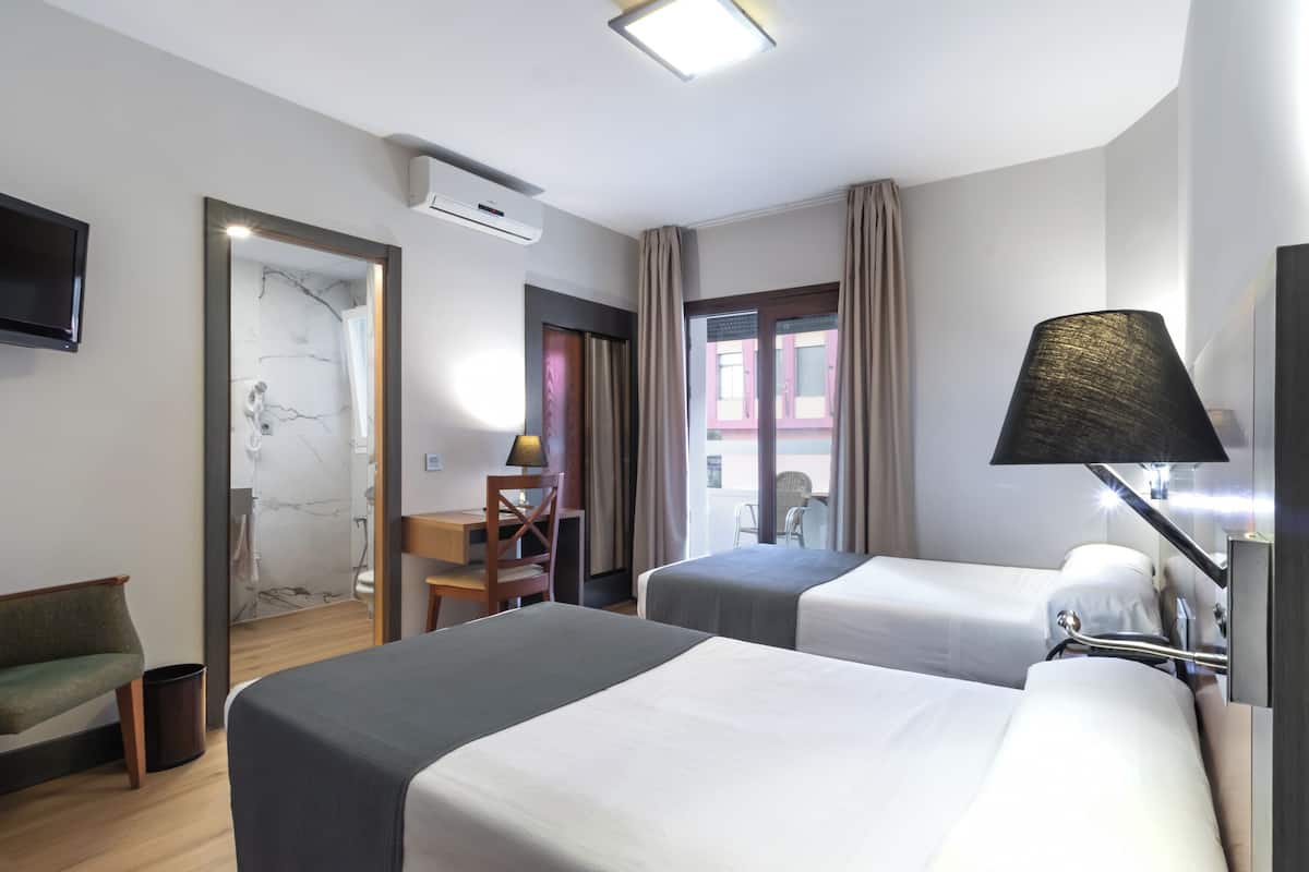 A hotel room with two beds, a desk, a chair, a TV, and a bathroom visible through an open door.