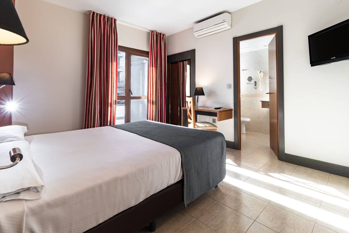 A hotel room with a bed, a desk, a chair, a TV, and a bathroom visible through an open door.