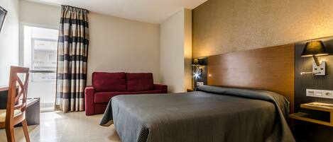 Una habitación de hotel con una cama grande, un sofá rojo, una mesita de noche de madera y un ventanal con cortinas.