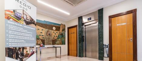 A hotel lobby with a reception desk, a large informational poster, a modern elevator, and a wooden door.