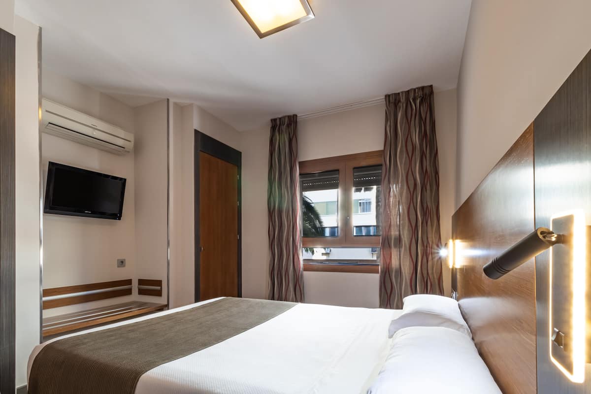 A hotel room with a bed, a flat-screen TV mounted on the wall, a window with curtains, and a wall-mounted light fixture.