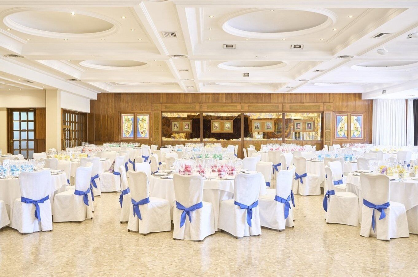 Un salón de banquetes con mesas redondas, sillas blancas adornadas con cintas azules y una pared de paneles de madera con vidrieras.