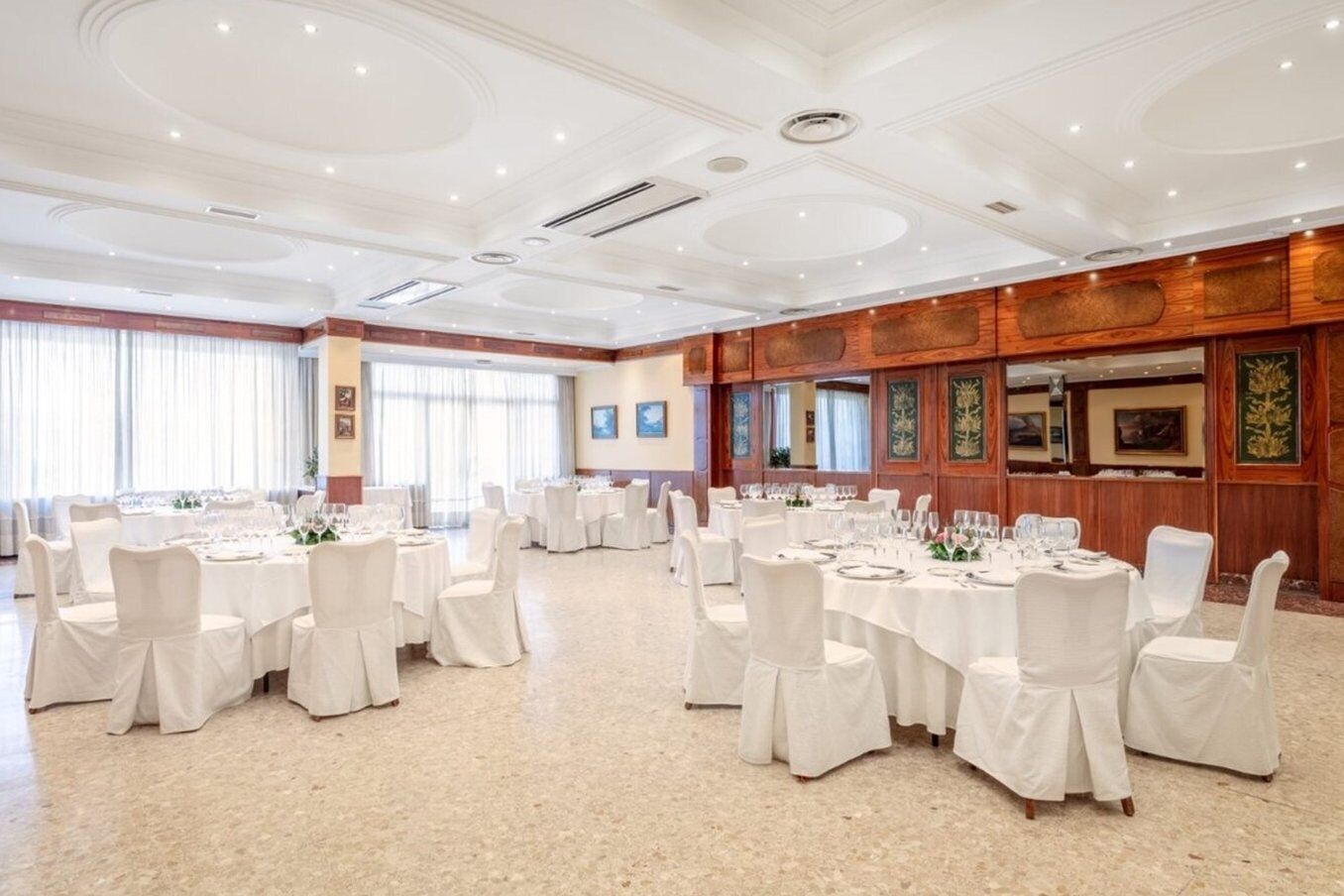Un amplio salón de banquetes con mesas redondas dispuestas para cenar, sillas blancas y suelo pulido.