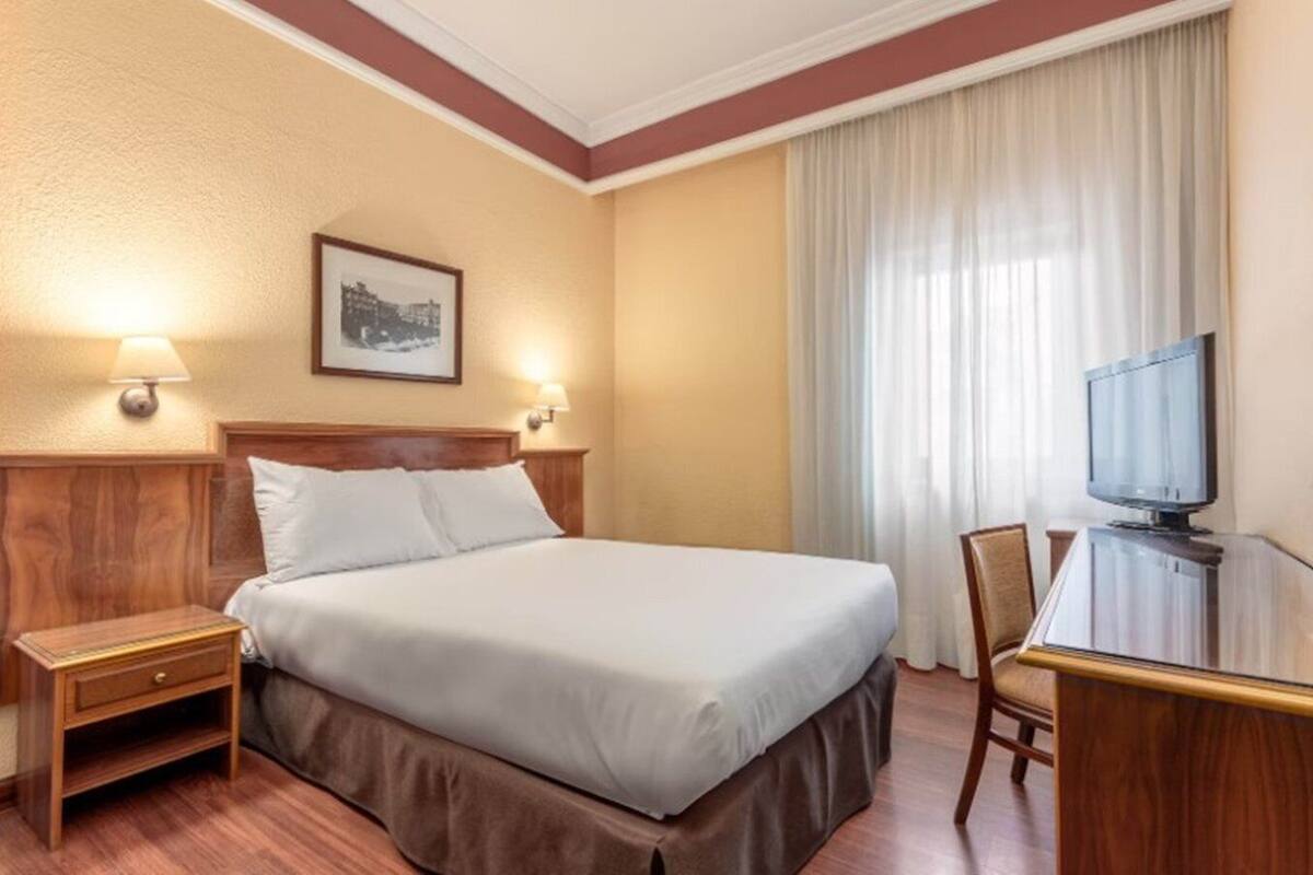 Una habitació d'hotel amb llit, taules de nit, escriptori amb cadira, televisió i finestra amb cortines.