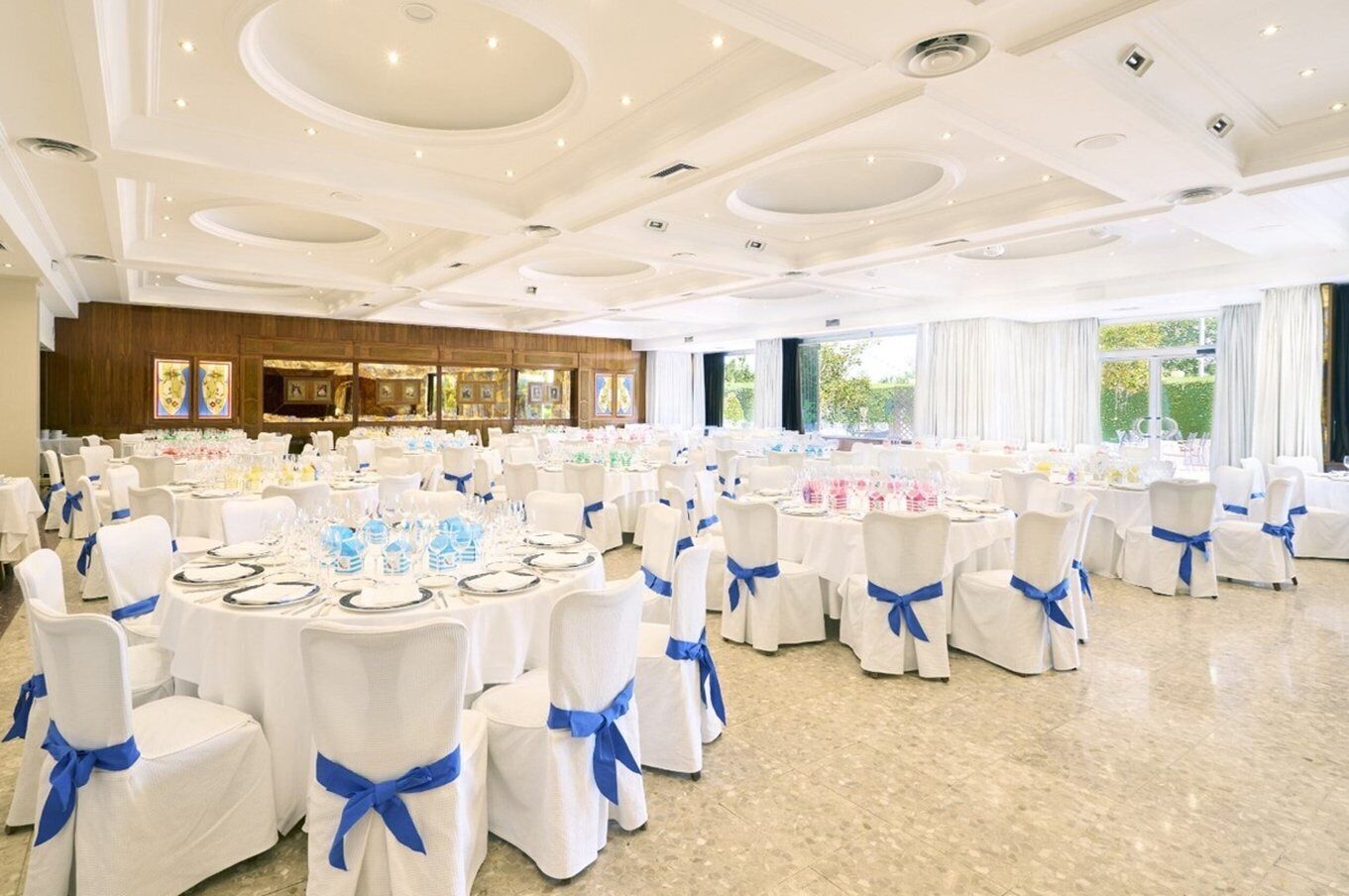 Un salón de banquetes con mesas redondas, sillas blancas con cintas azules y un techo alto con iluminación empotrada.