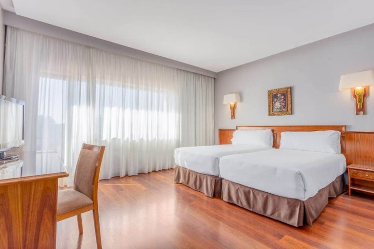 Habitació d'hotel amb llit de matrimoni, escriptori amb cadira, televisió i finestra gran amb cortines transparents.