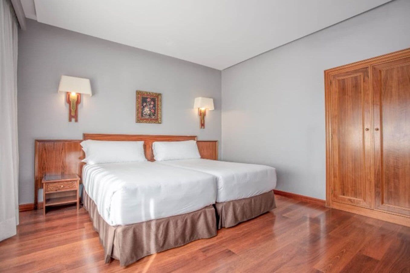 A double bed with a wooden headboard and skirt, two bedside lamps, a nightstand, a large wardrobe, and a framed picture on the wall.