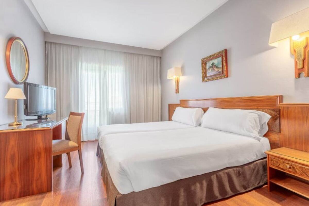 Habitació d'hotel amb un llit gran, un escriptori amb televisió, una cadira i una finestra amb cortines.
