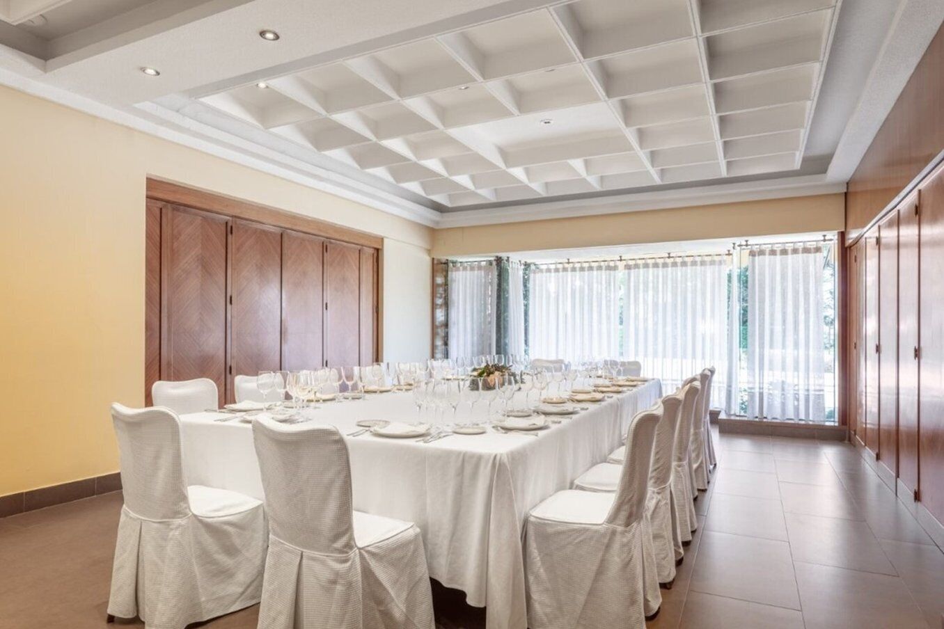 A conference room with a long table set for a meeting, large windows with curtains, and wooden paneling on the walls.