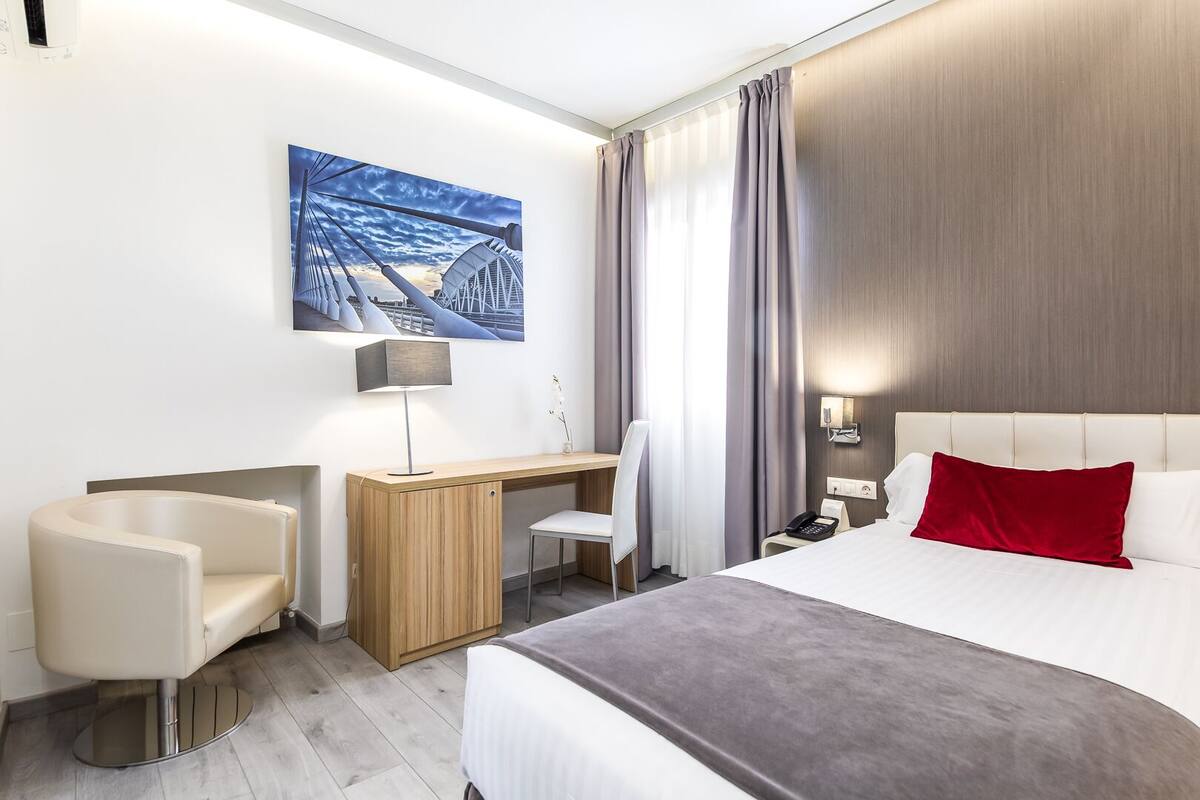 A hotel room with a bed, a desk, a chair, a lamp, a phone, and a painting on the wall.