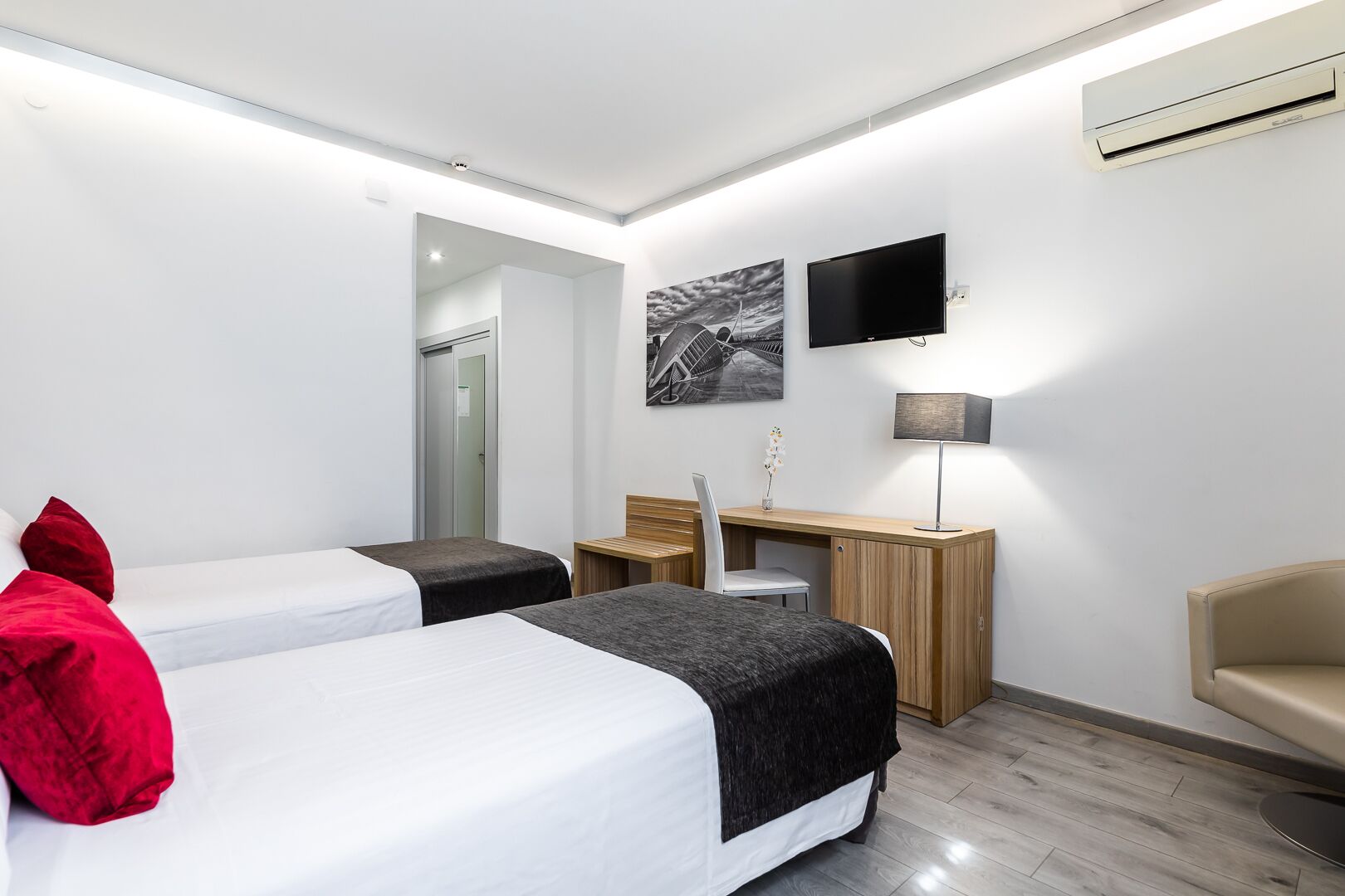 Habitación de hotel moderna con dos camas, un escritorio, una silla, un televisor y aire acondicionado instalado en la pared.