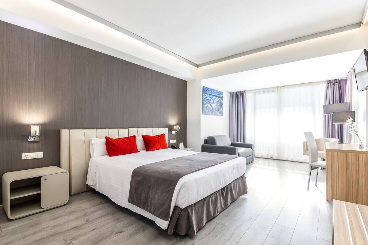 A modern hotel room with a large bed, bedside tables, a desk, and a view of the city.