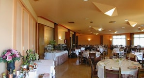 Un amplio comedor con varias mesas redondas dispuestas para comer, una zona de bufé con comida y bebidas, y un gran ventanal que proporciona luz natural.