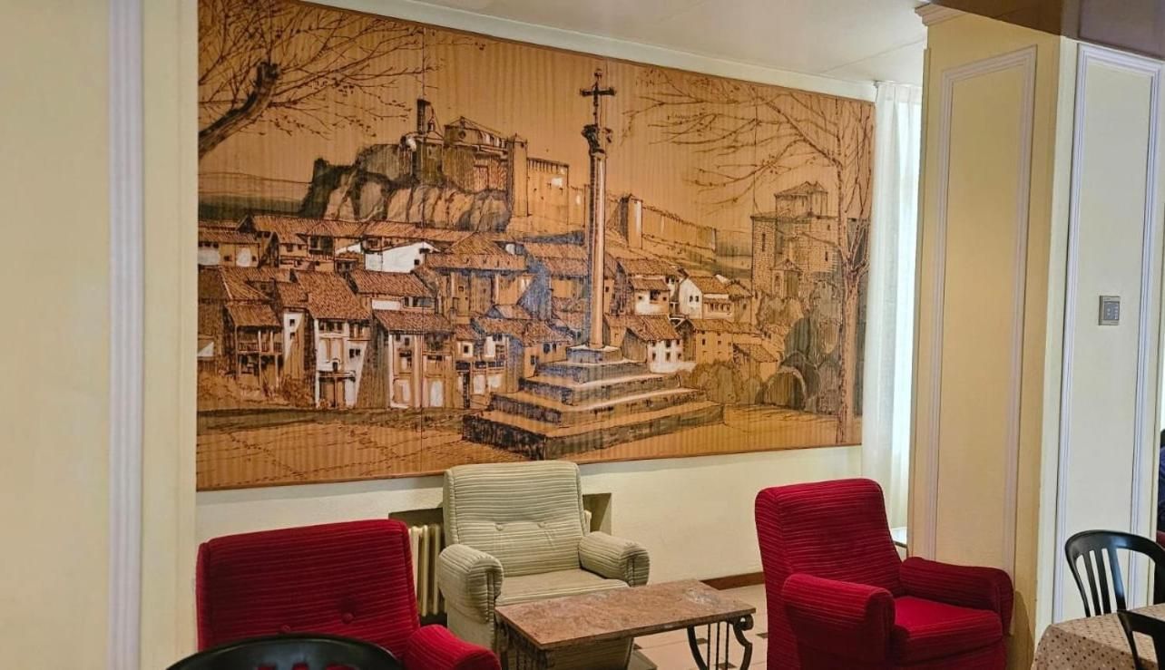 Una habitación con un gran mural que representa una escena urbana, una pequeña mesa y dos sillas rojas.