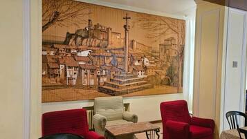 Una habitación con un gran mural que representa una escena urbana, una pequeña mesa y dos sillas rojas.