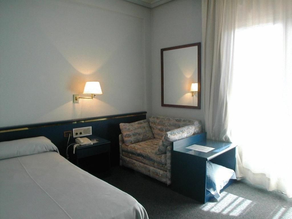 A hotel room with a bed, a chair, a desk, a lamp, a telephone, and a mirror.