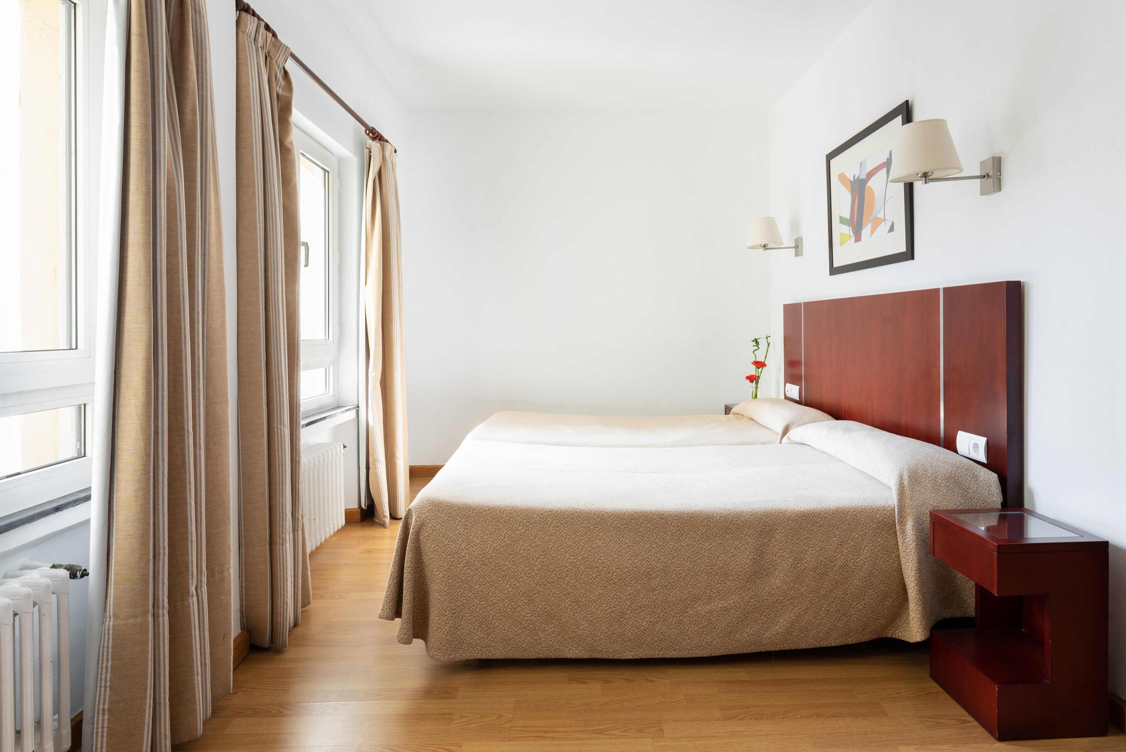 A hotel room with a large bed, a wooden nightstand, a framed picture on the wall, and a window with curtains.