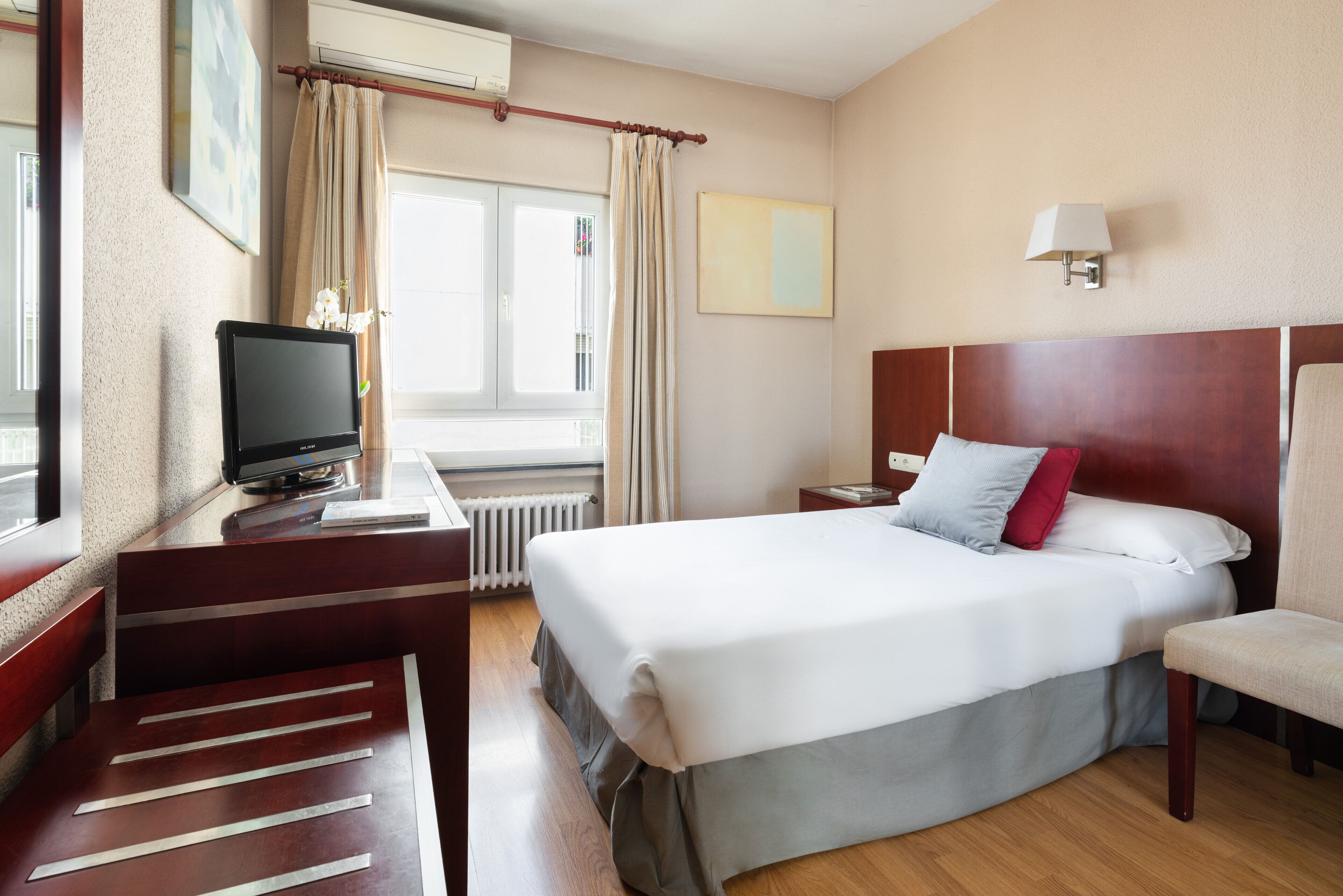 A hotel room with a bed, a desk with a television, a chair, a window with curtains, and a wall-mounted lamp.