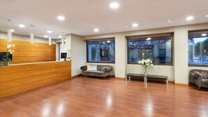 A reception area with a wooden reception desk, a grey sofa, a bench, and large windows with a view of the street. - Crisol Riosol (León)
