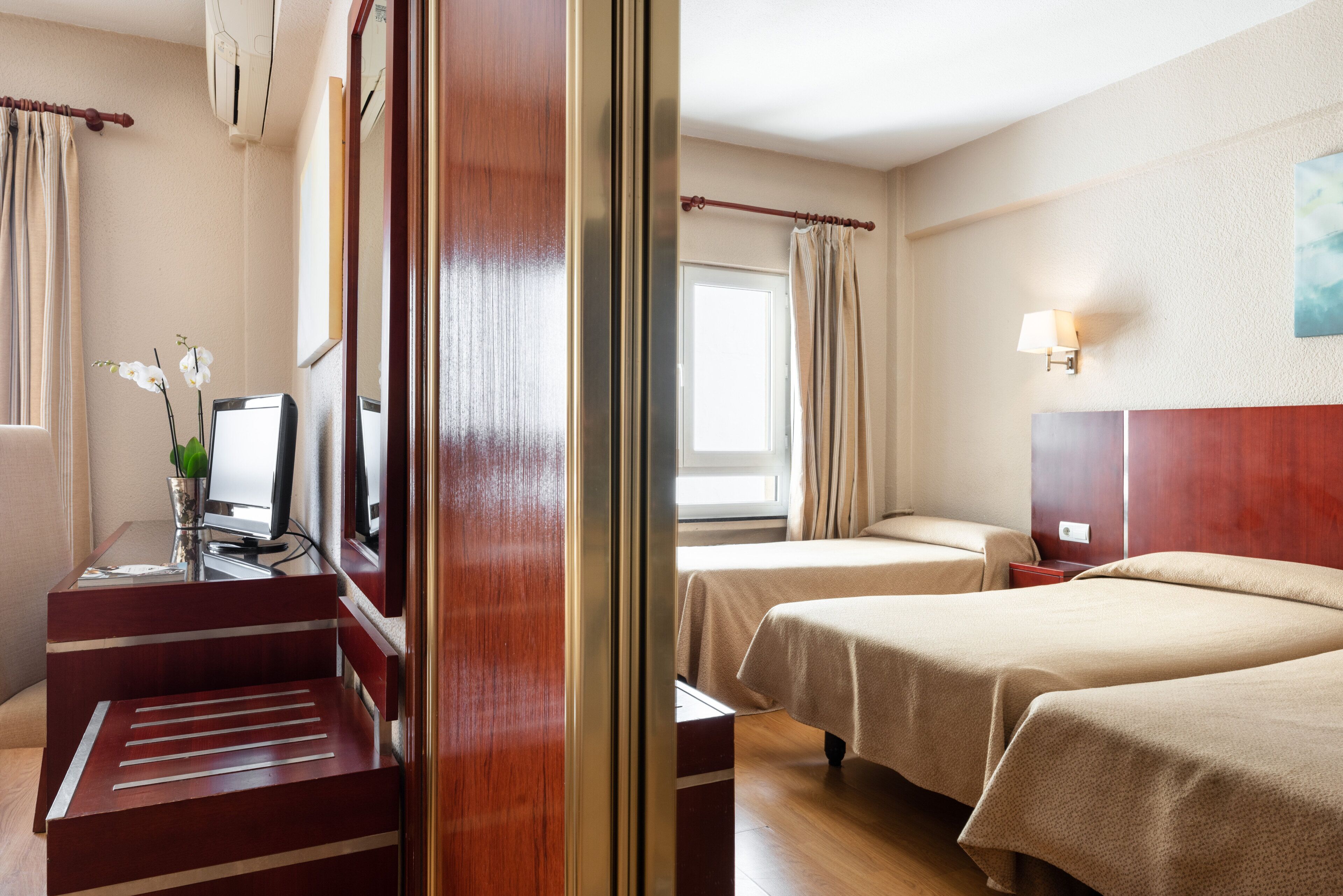 Habitación de hotel con dos camas, un escritorio con computadora, una lámpara y un ventanal con cortinas.