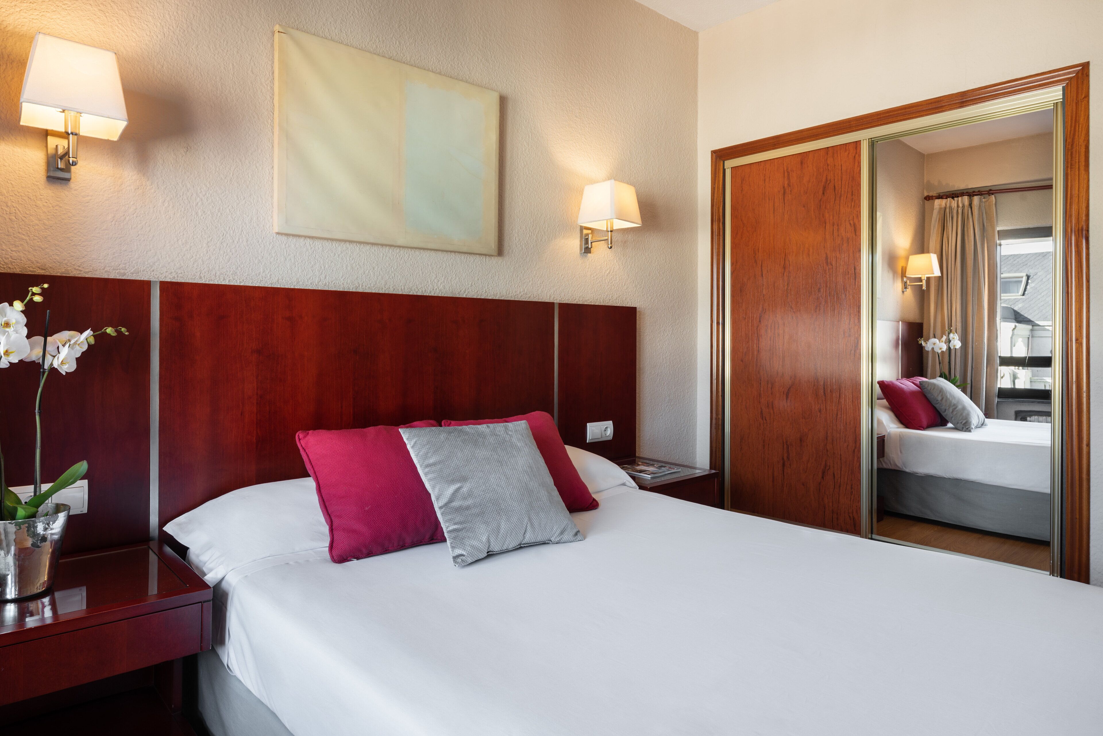Ein Hotelzimmer mit einem großen Bett, einem Holz-Kopfteil, Nachttischlampen, einem Spiegel und einem Nachttisch mit Blumen.